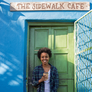 Woman holding biodegradable coffee cup against bright blue wall and under a 'Sidewalk Cafe' sign