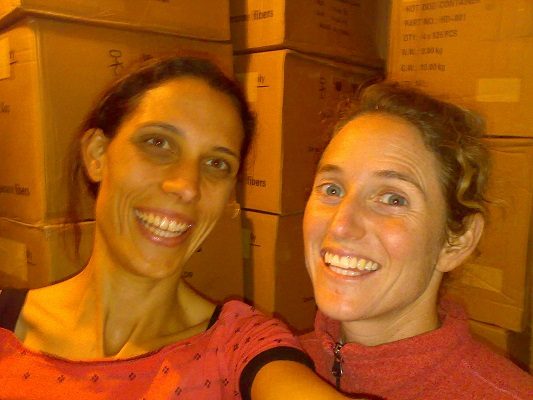 Catherine with a friend, both smiling happily in front of boxes of stock