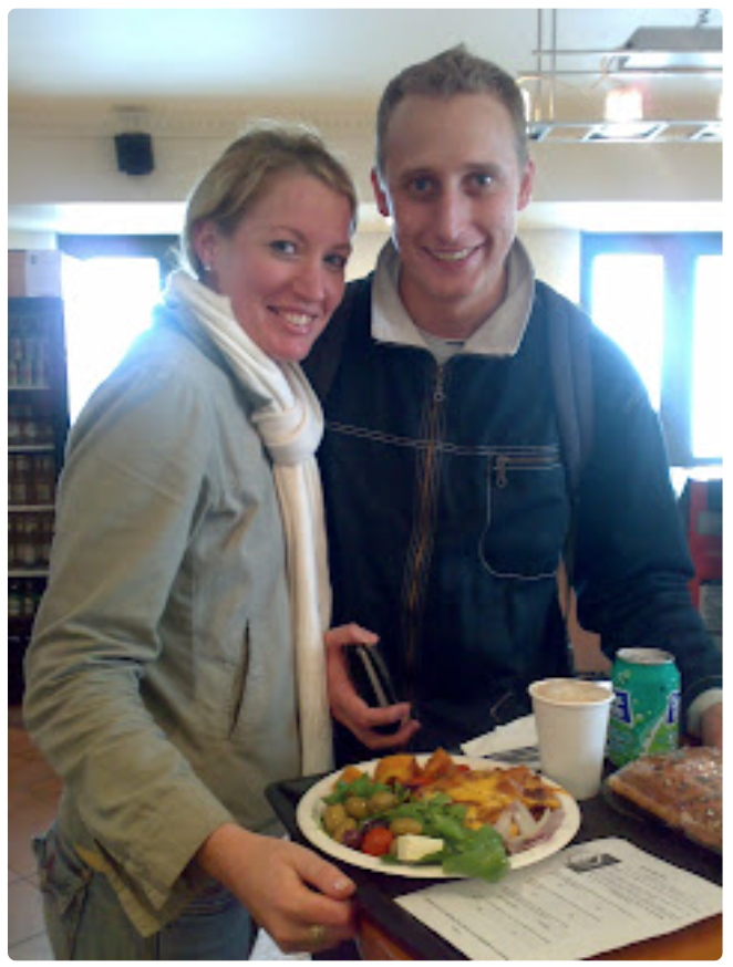 A smiling tourist couple standing at table with food on a bagasse plate and coffee in a paper cup